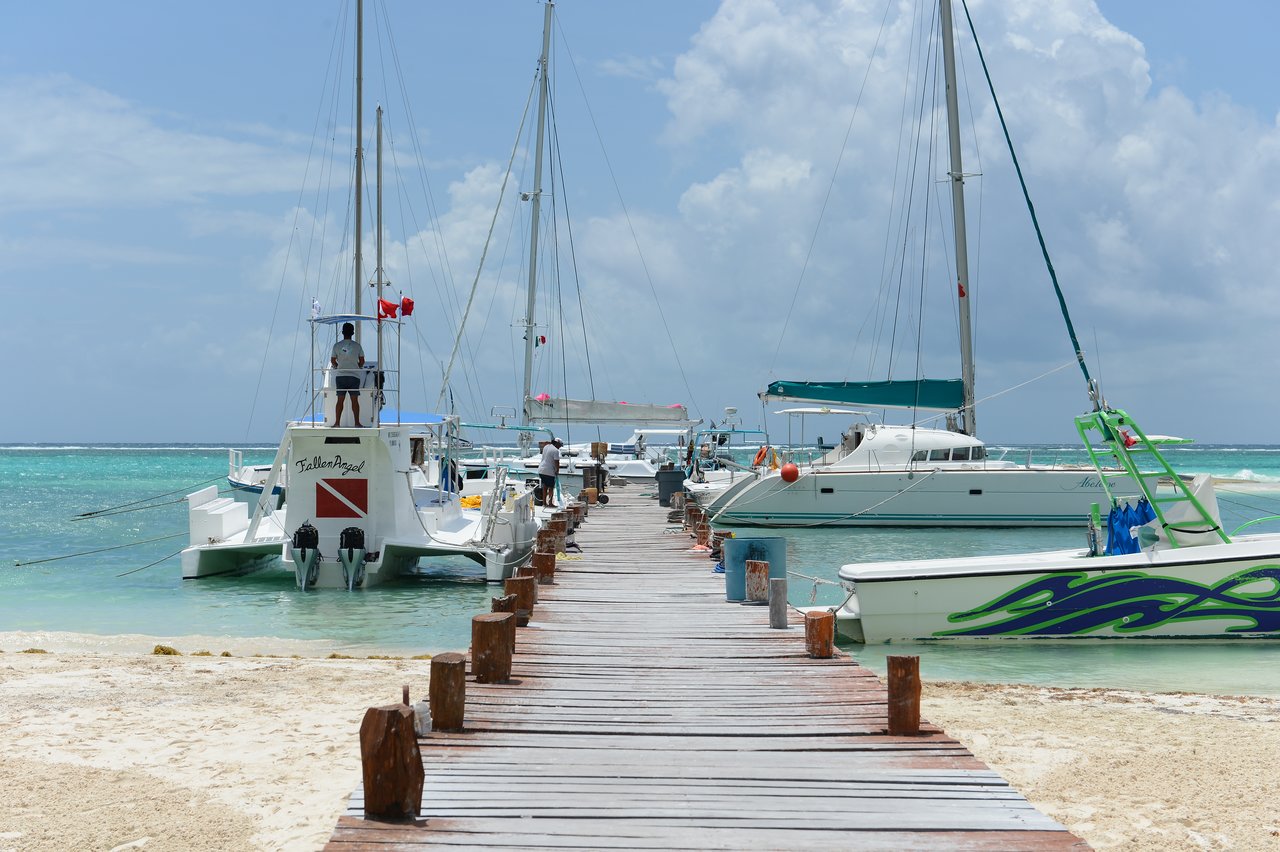 A wooden pier extends into the ocean, with several boats docked and people walking or standing on board.