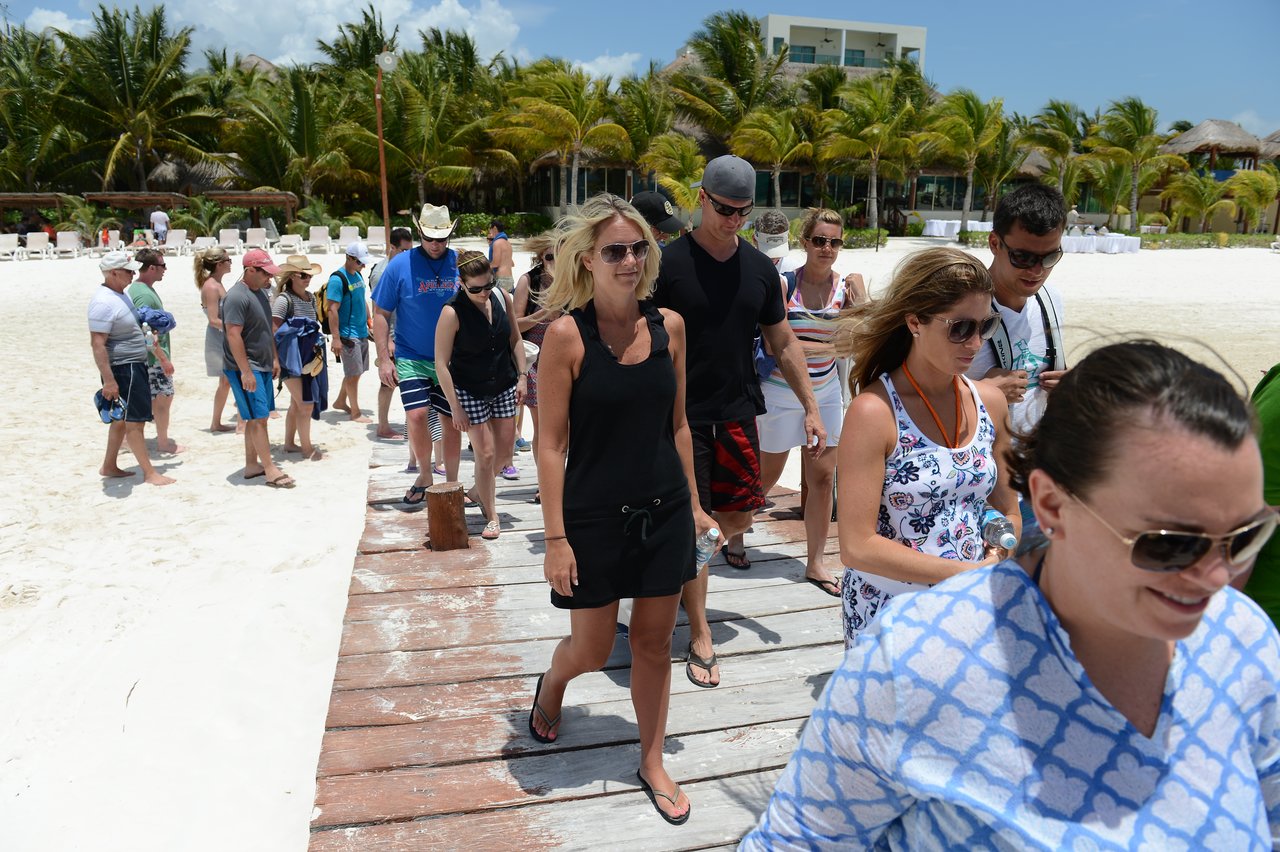 A group of people walks along a wooden pier toward the beach, dressed in casual summer clothing and sunglasses.