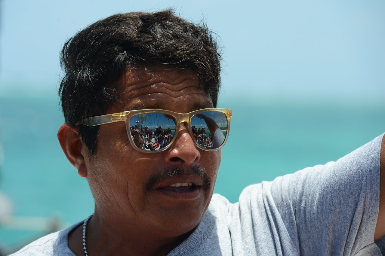 A man wearing sunglasses speaks, with a reflection of a group of people on a boat visible in his lenses.