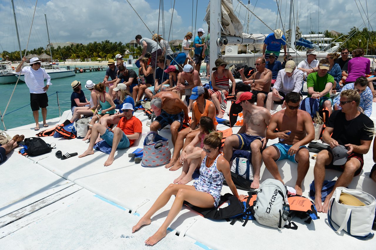 A group of people sits on a catamaran deck, wearing summer clothes and hats, preparing for a boat trip.