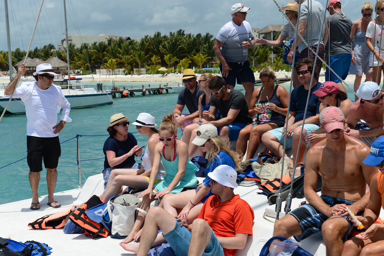 A group of people relax on a boat, wearing hats and sunglasses, enjoying drinks near the shore.
