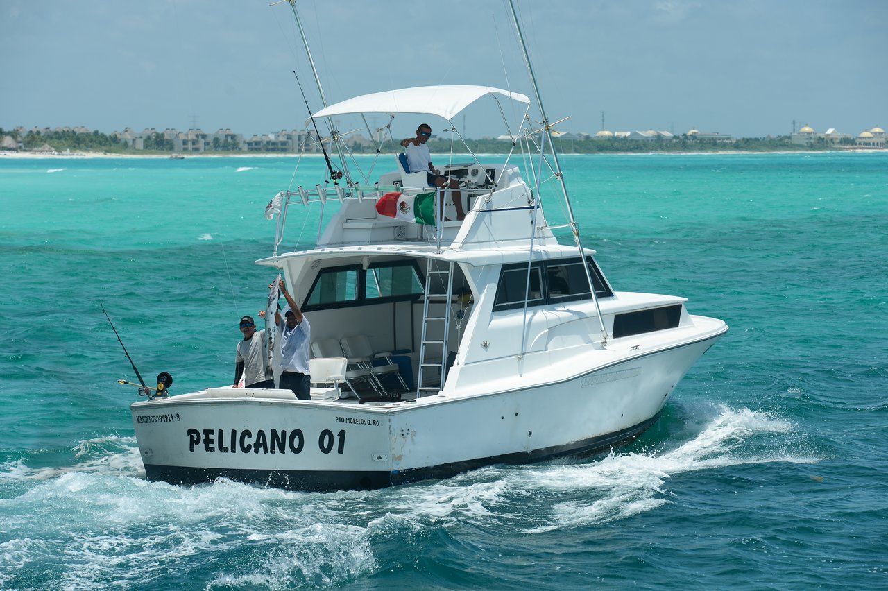 A fishing boat named "Pelicano 01" moves through turquoise water, with people on board holding up a catch.