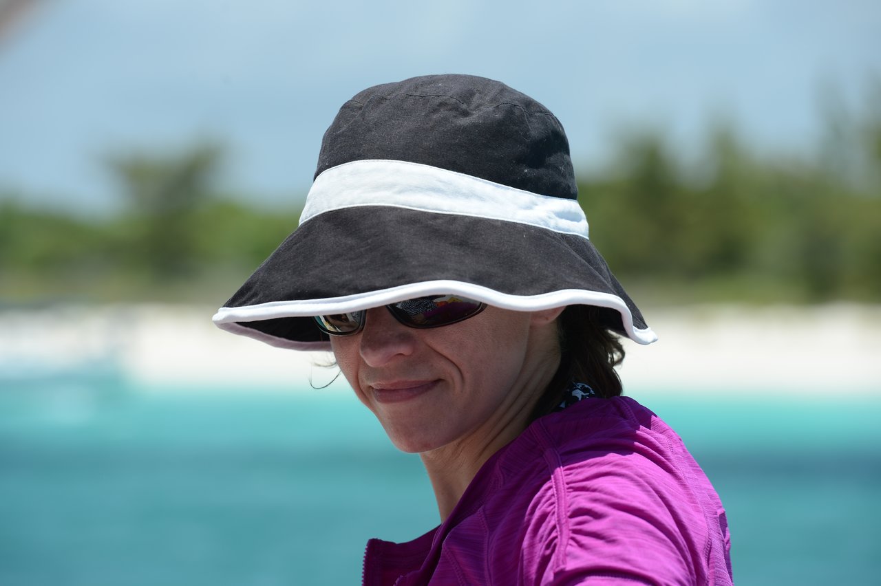 A woman wearing a sun hat and sunglasses smiles while outdoors near the beach.