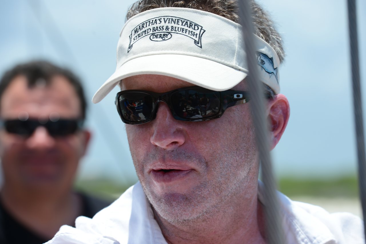A man wearing sunglasses and a visor looks ahead, with another person slightly blurred in the background.