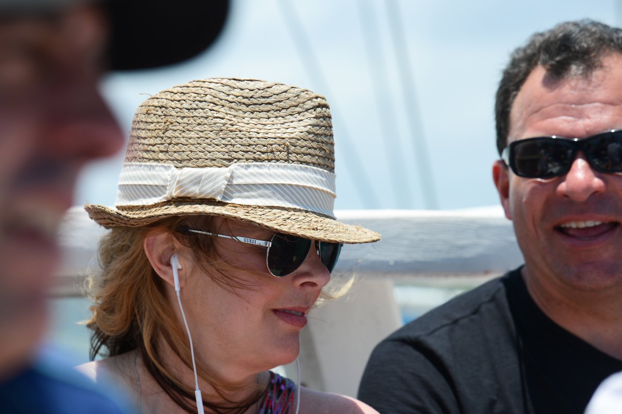 A woman wearing a straw hat and sunglasses listens to earphones while sitting next to a man in sunglasses.