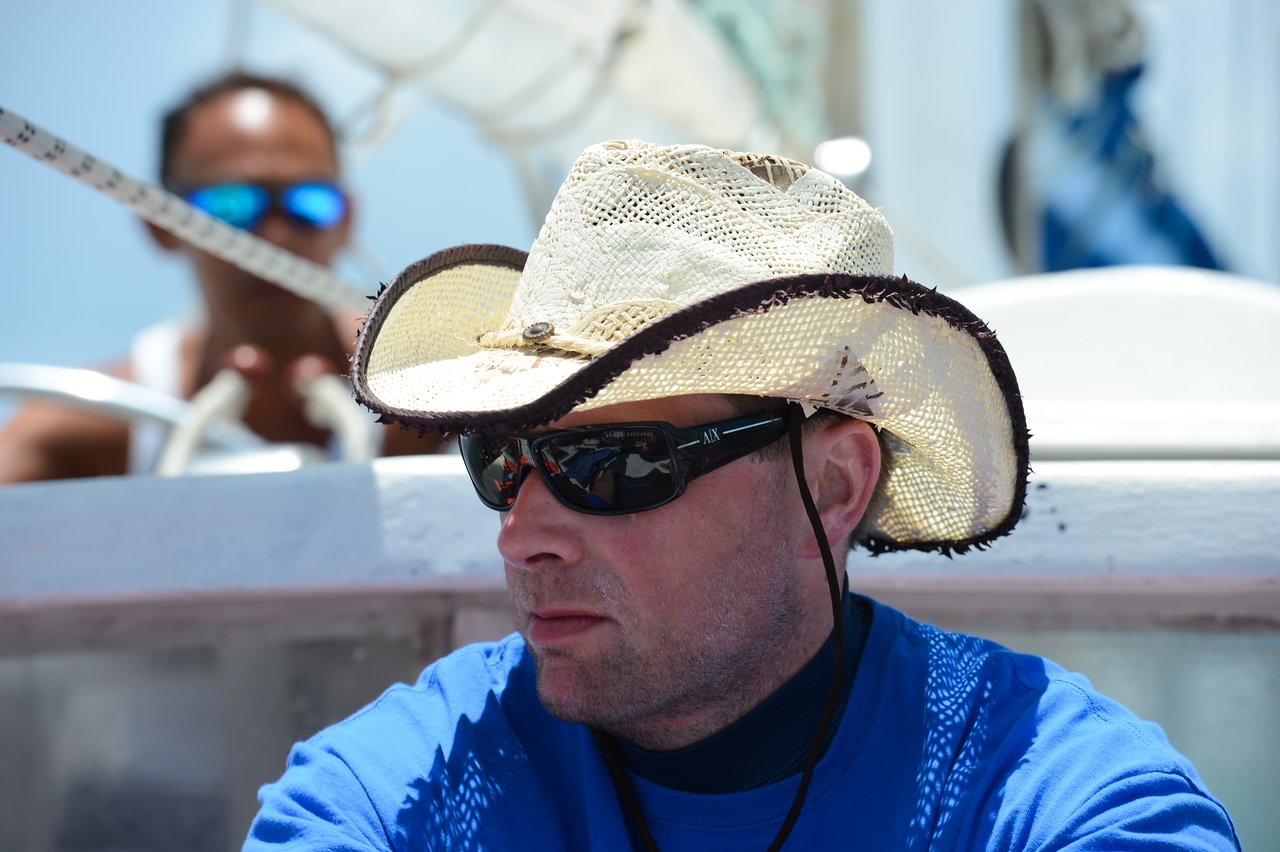 A man wearing a straw hat and sunglasses sits on a boat, looking to the side.