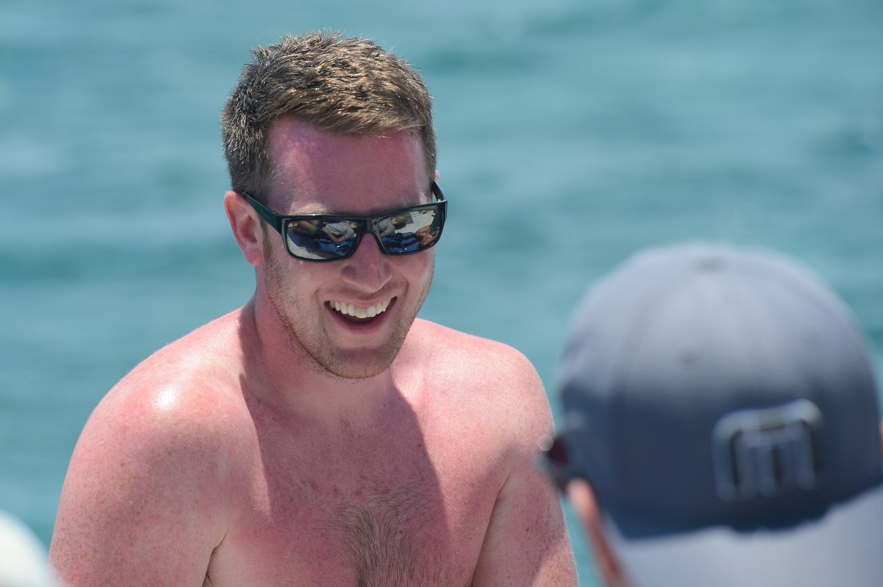 A smiling, shirtless man wearing sunglasses is outdoors near the water, talking to someone wearing a cap.