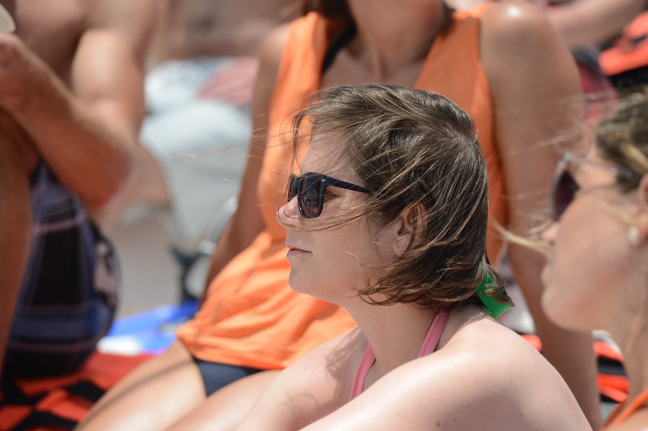 A woman wearing sunglasses sits among a group of people, looking ahead in a sunny outdoor setting.