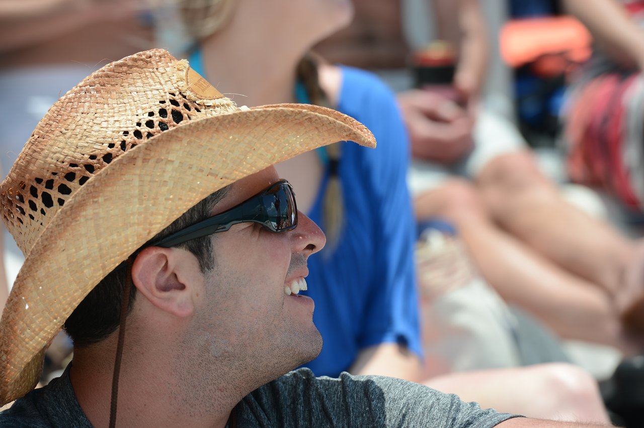 A man wearing a straw hat and sunglasses smiles while sitting among a group of people outdoors.