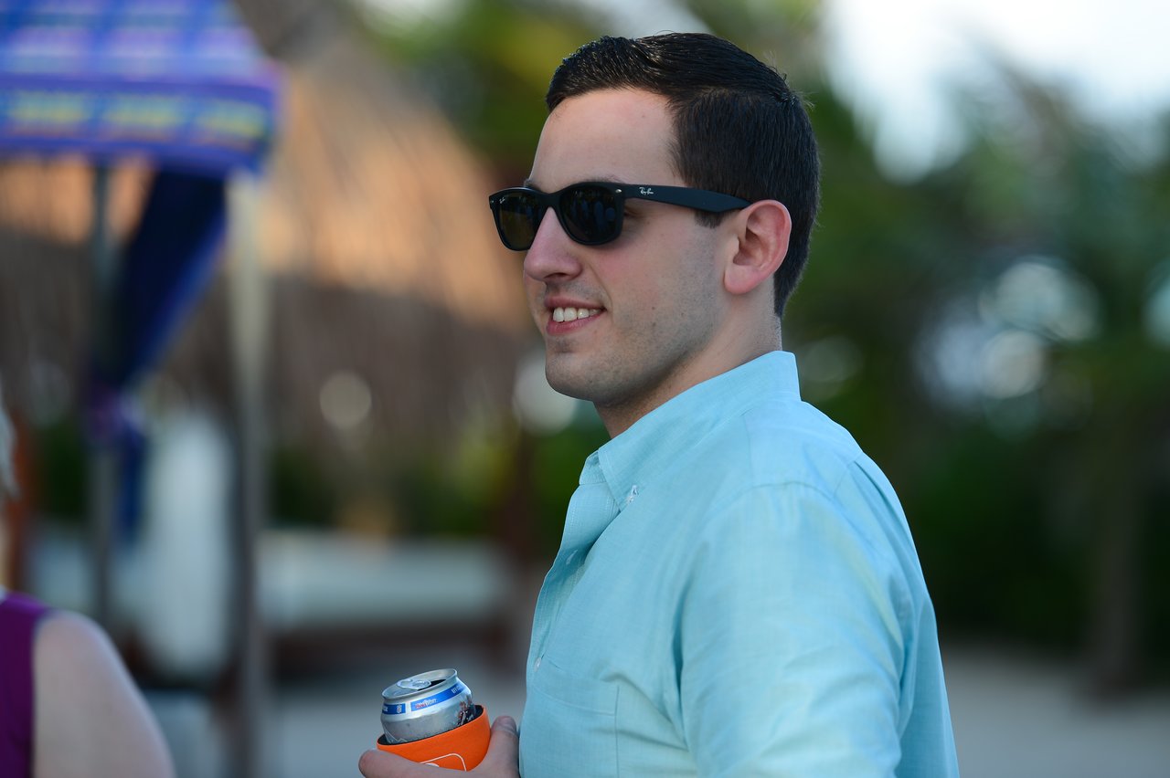A man wearing sunglasses and a light blue shirt smiles while holding a canned drink in Cancun.