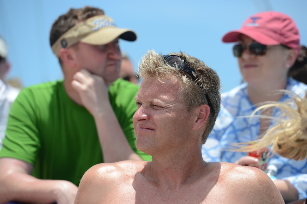 A man squints in the sunlight while sitting shirtless outdoors, with two people in the background wearing hats and sunglasses.