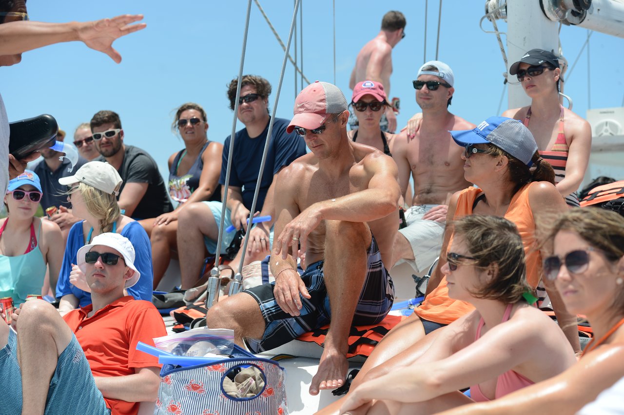 A group of people in swimwear sit on a boat, listening to a person speaking under the bright sun.