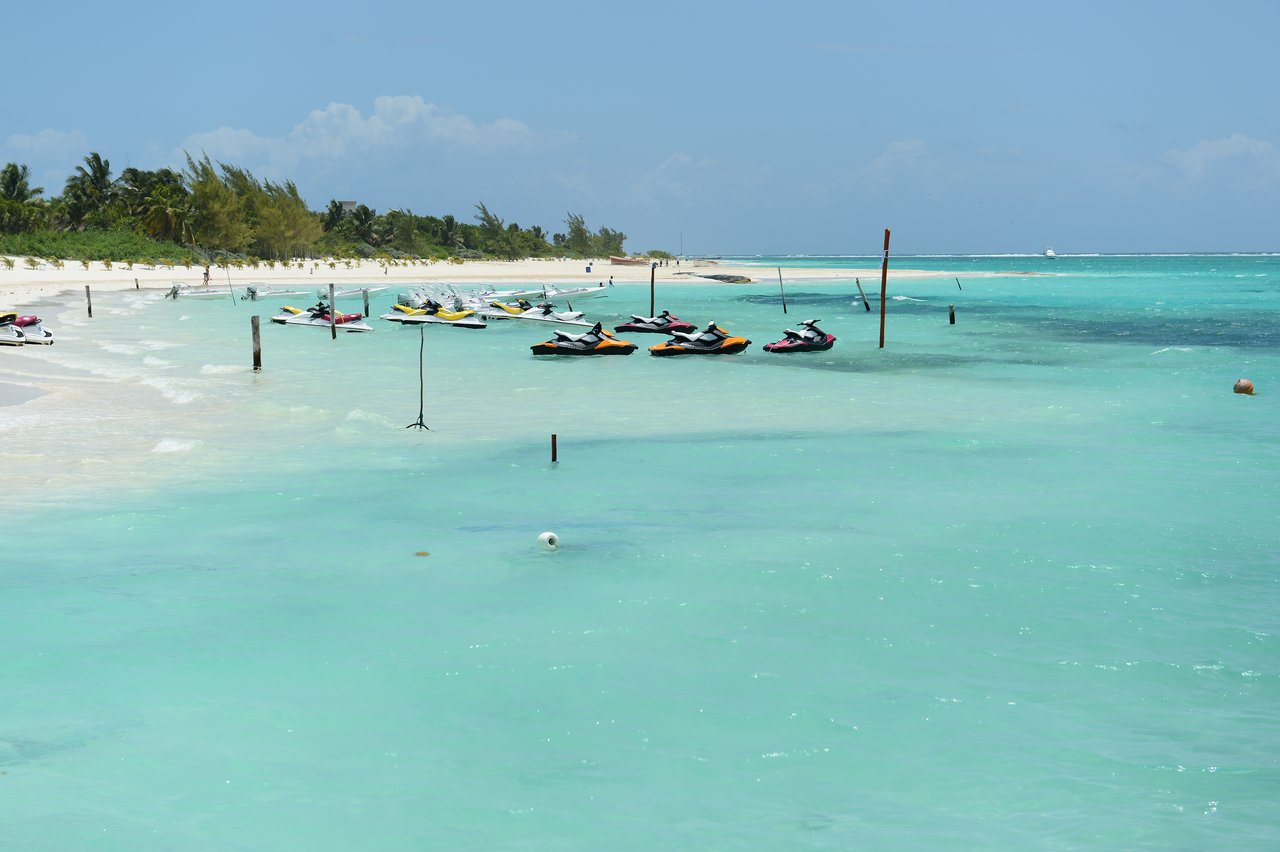 Jet skis floating on clear turquoise water near a sandy beach with palm trees in the background.