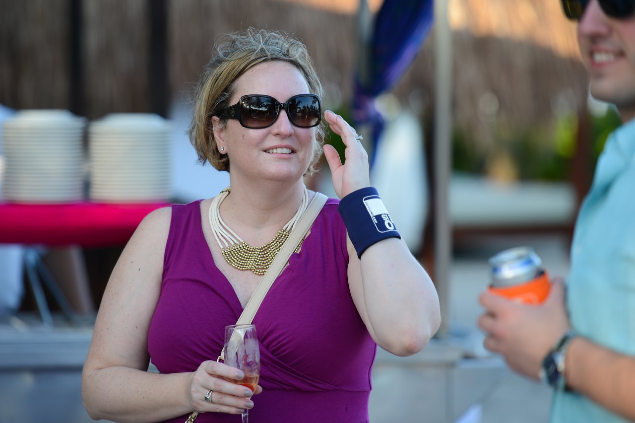 A woman in sunglasses and a purple dress holds a drink and adjusts her hair at an outdoor gathering.