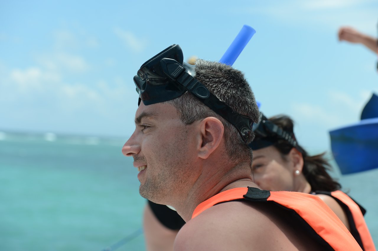 A man wearing a snorkel mask and life vest smiles while looking at the ocean, with others nearby.