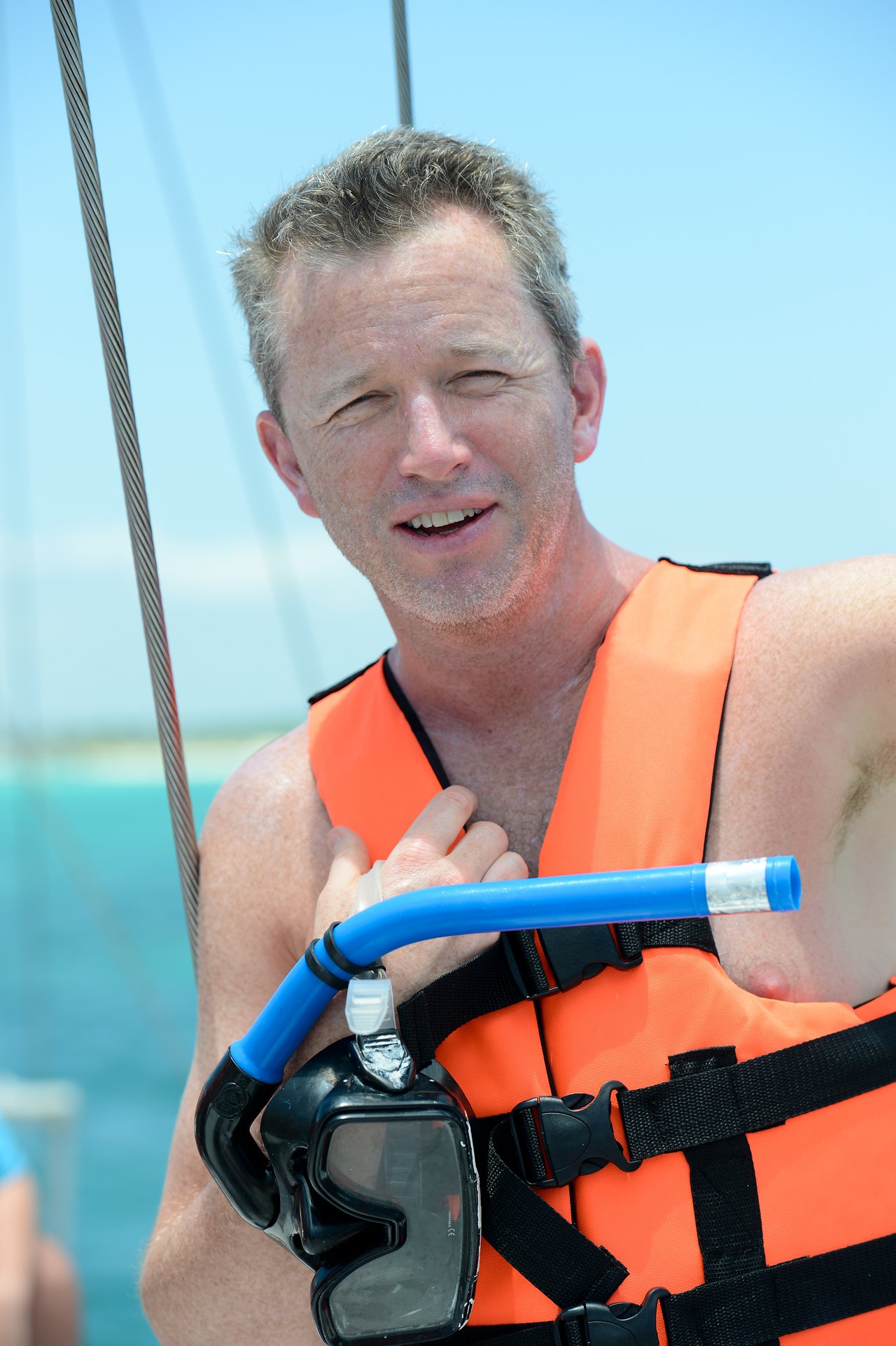 A man wearing an orange life vest holds a snorkeling mask and tube while standing on a boat.