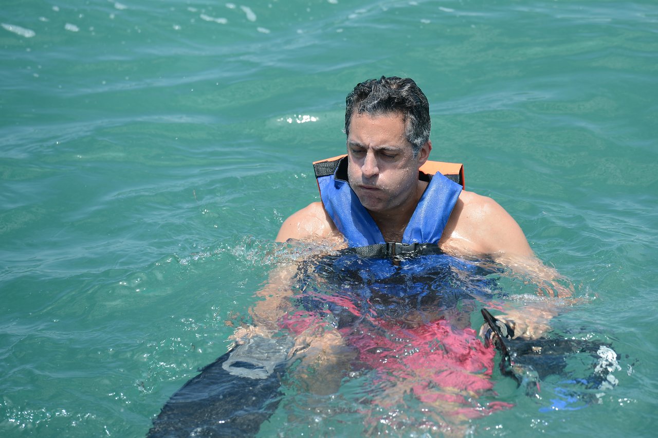 A man wearing a life jacket floats in the water, holding swim fins and puffing his cheeks.