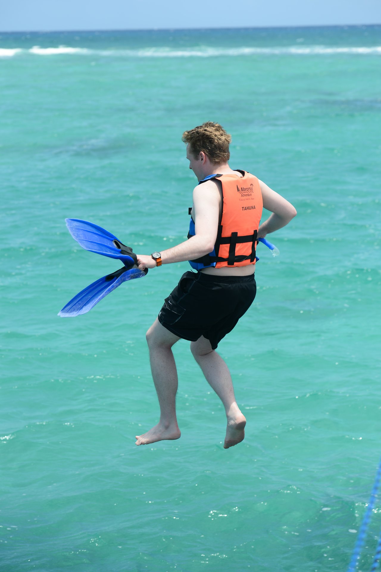 A person wearing a life vest and holding flippers jumps into the clear turquoise water.