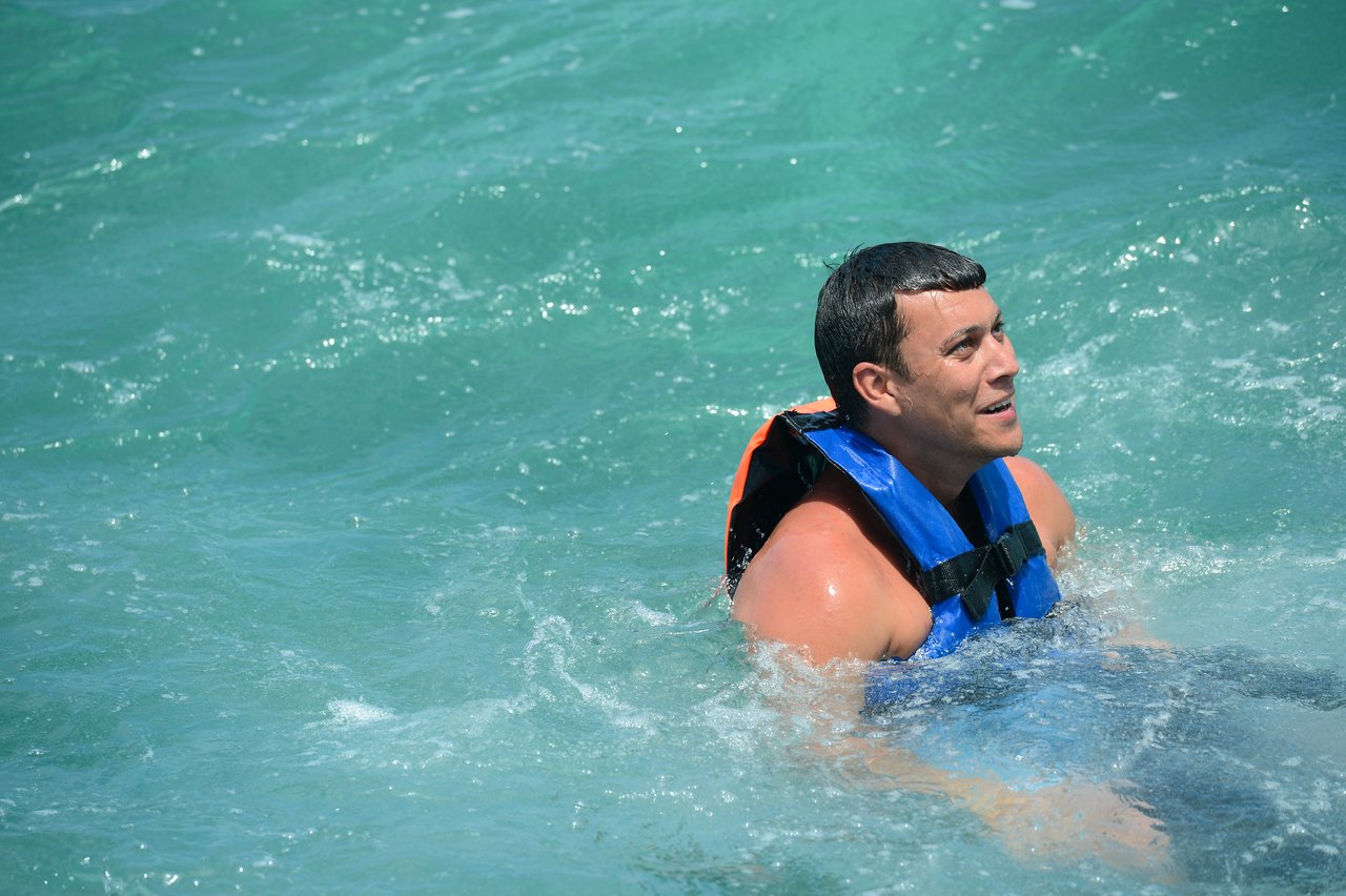 A man wearing a blue life jacket swims in the ocean, smiling and looking ahead.