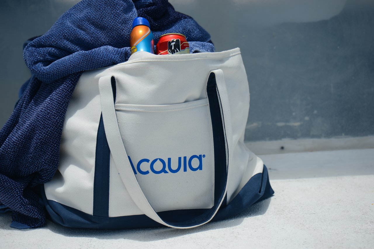 A beach bag with a towel, sunscreen, and a soda can resting on a concrete surface.