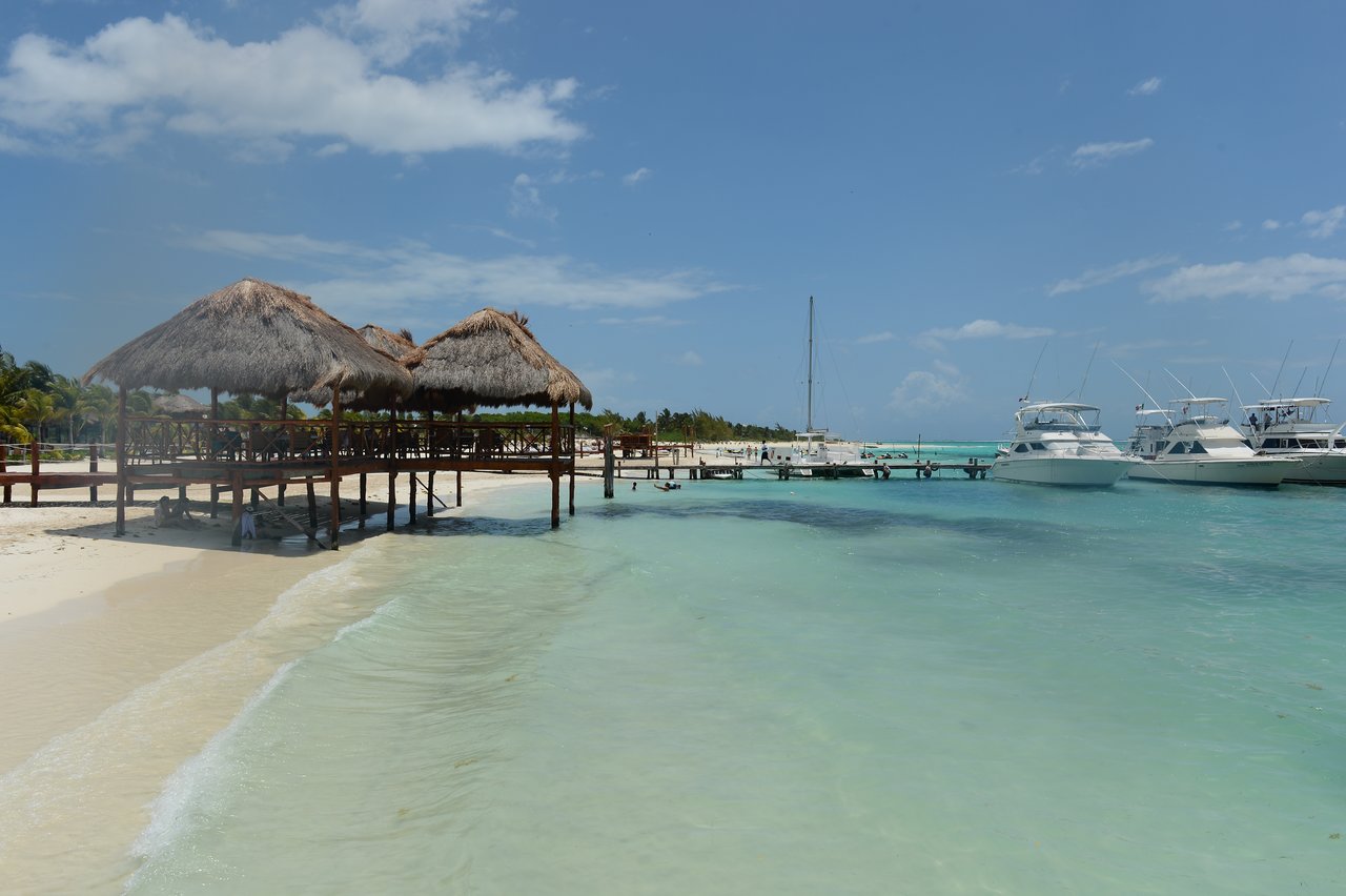Thatched-roof huts on a wooden pier extend over clear water, with boats docked nearby under a sunny sky.