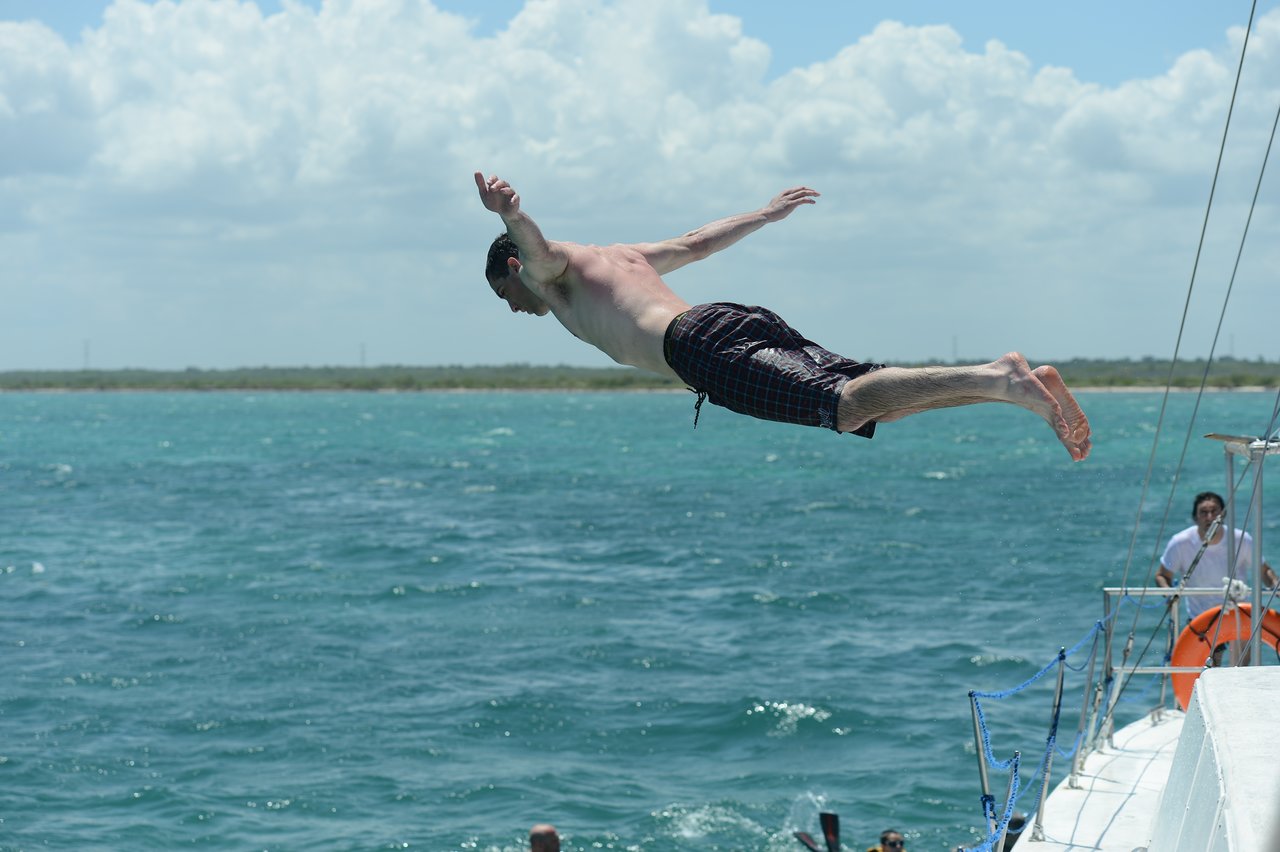A man in swim trunks jumps off a boat into the ocean, arms outstretched.