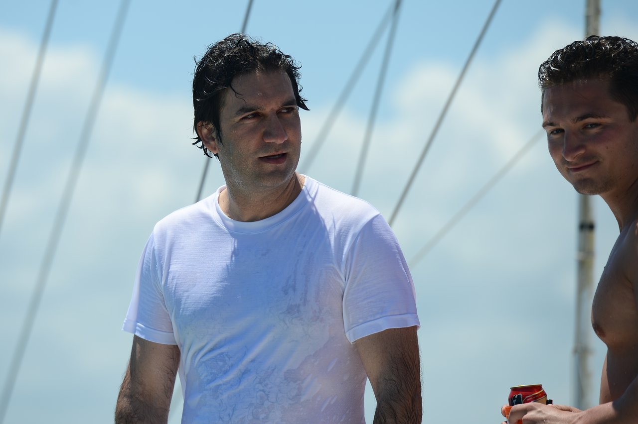 Two men on a boat, one in a wet white shirt and the other holding a drink, under a sunny sky.
