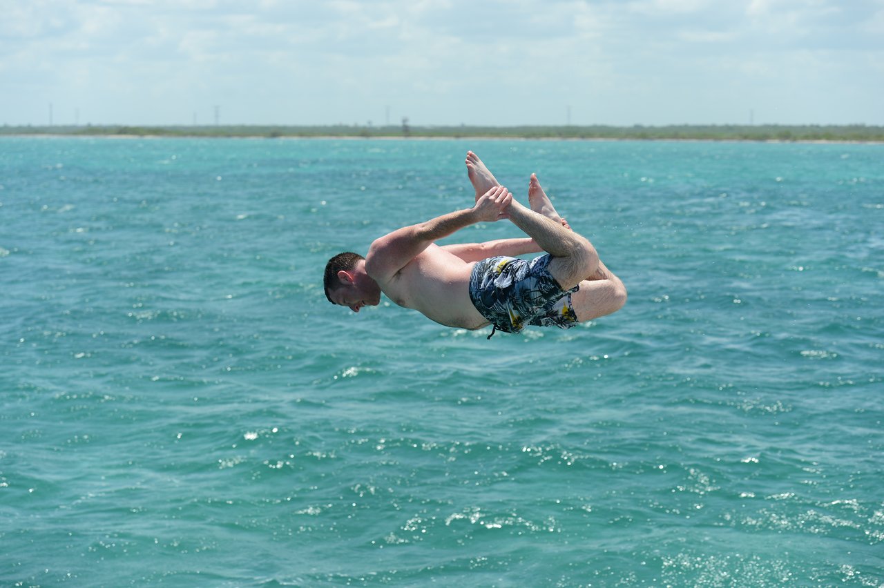 A man in swim trunks flips mid-air above the ocean, preparing to dive into the water.