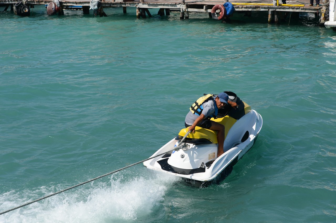 A man wearing a life jacket and cap rides a jet ski, holding onto a rope near a dock.