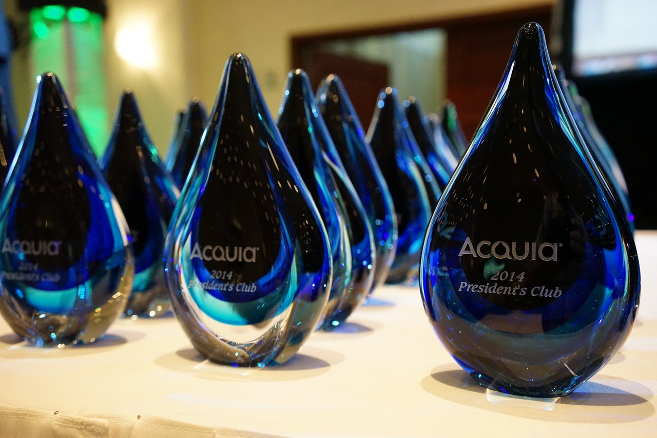 A table displays multiple blue glass awards labeled "Acquia 2014 President's Club" at an award party.