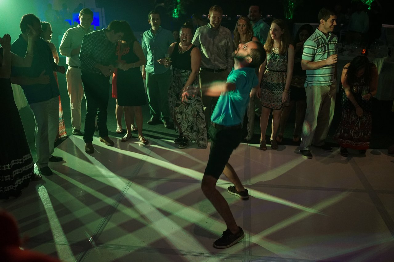 A man in a blue shirt dances energetically on a lit dance floor while a crowd watches and cheers.