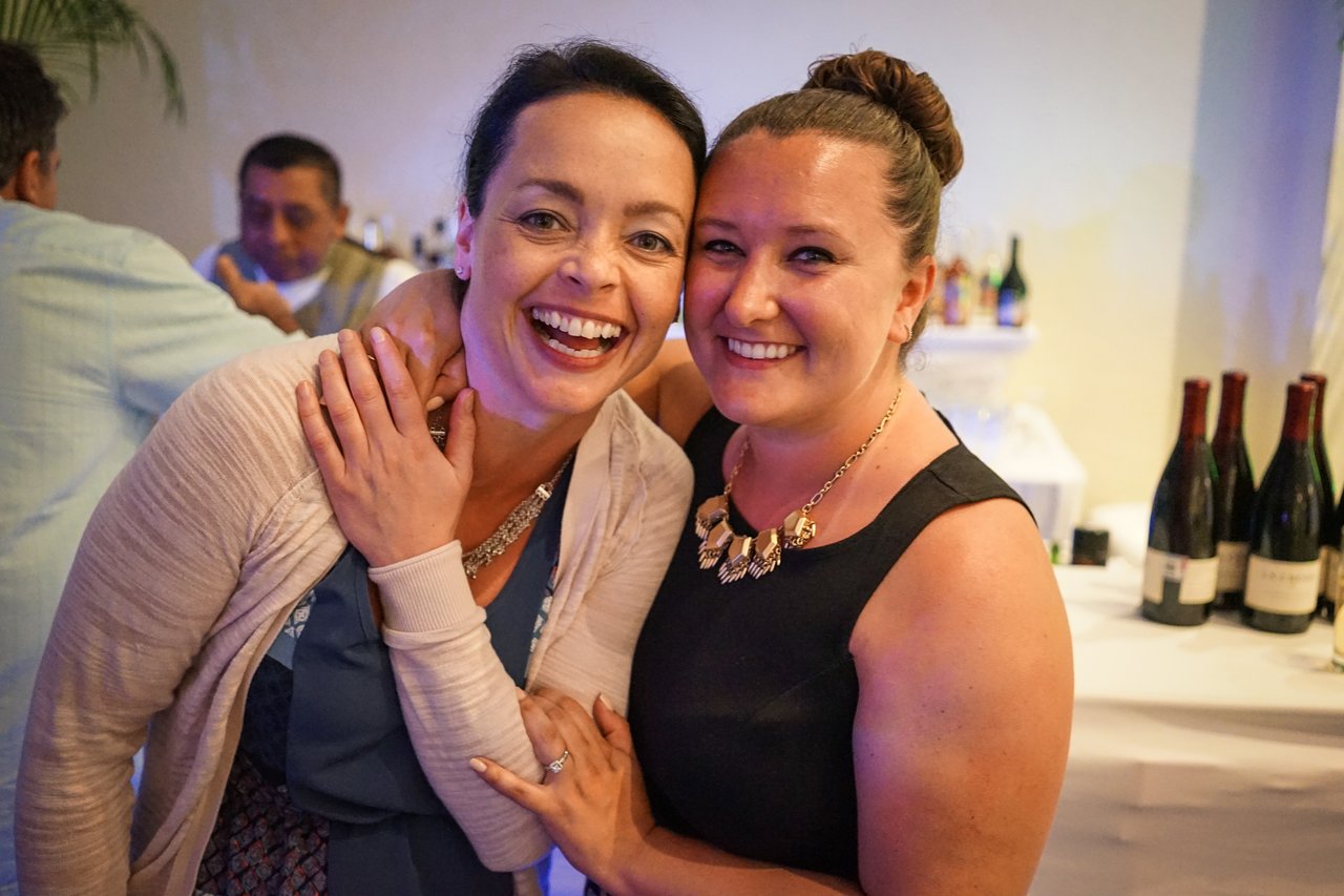 Two smiling women pose together at an award party, with drinks and wine bottles visible in the background.