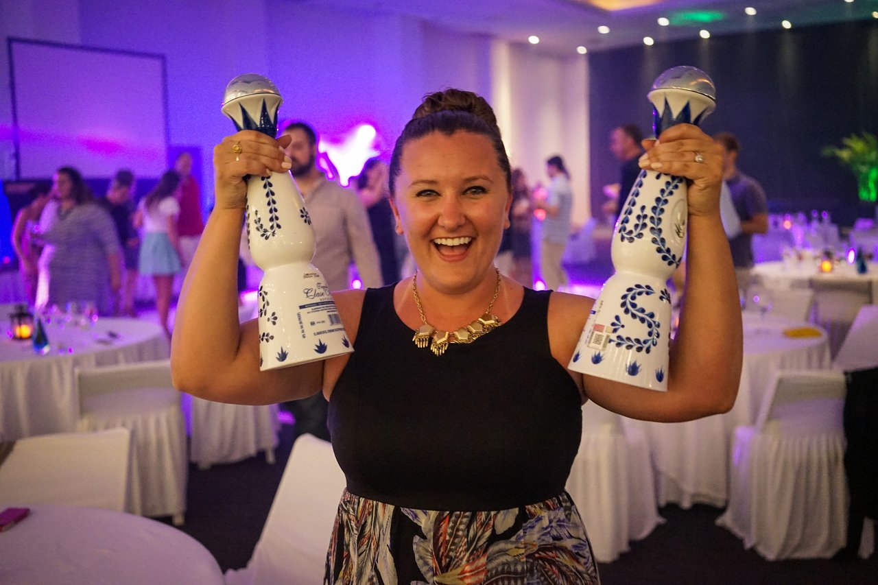 A smiling woman holds two decorative bottles at an award party, surrounded by people in a festive setting.