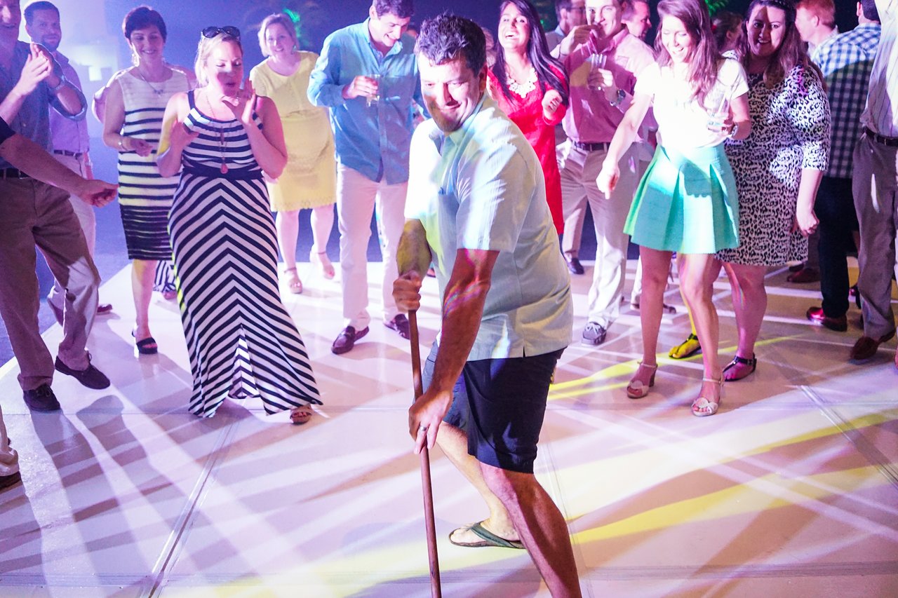 A man dances with a stick on a lit-up floor while a crowd watches and cheers at a party.