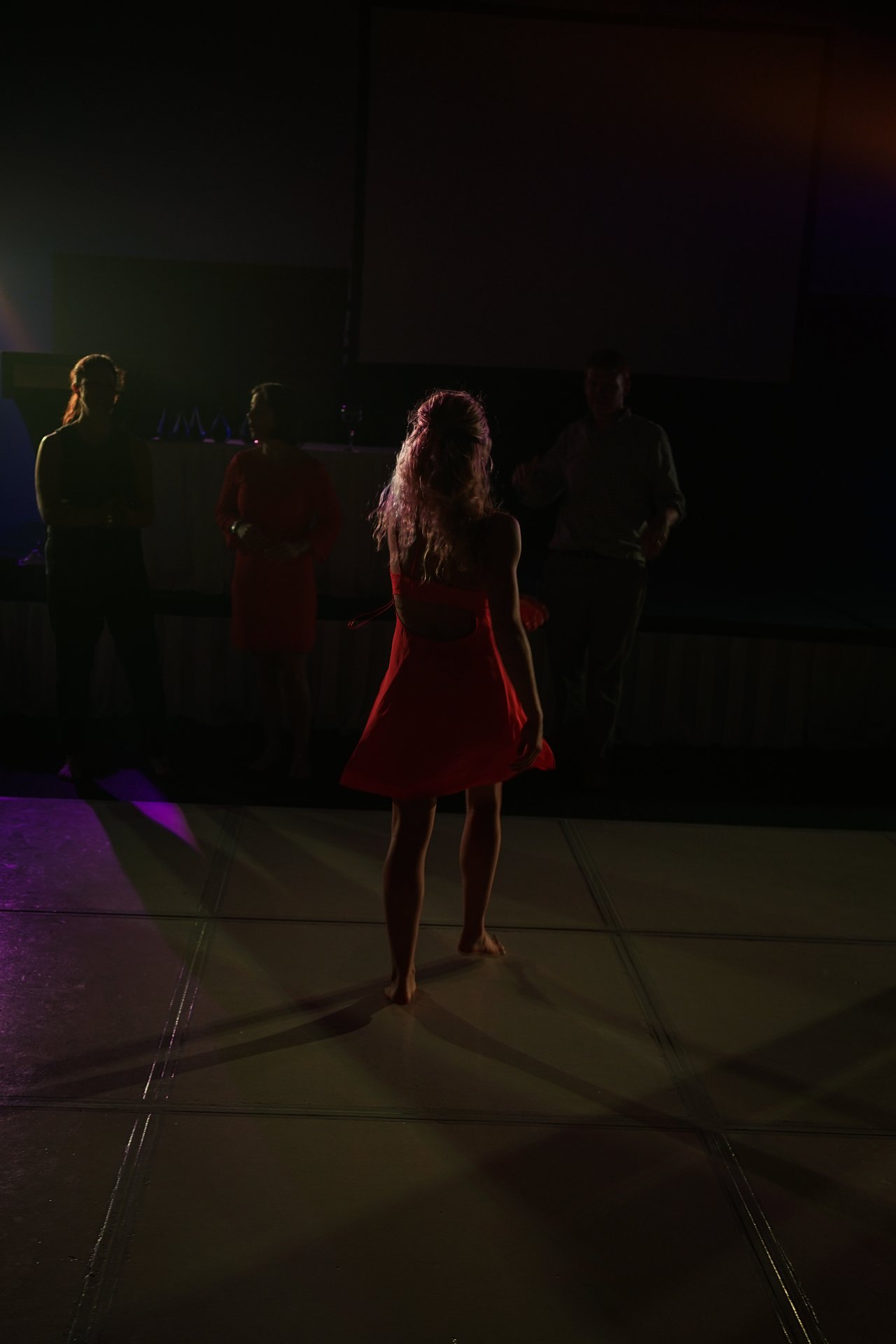 A woman in a red dress stands barefoot on a dance floor, facing a group of people in dim lighting.