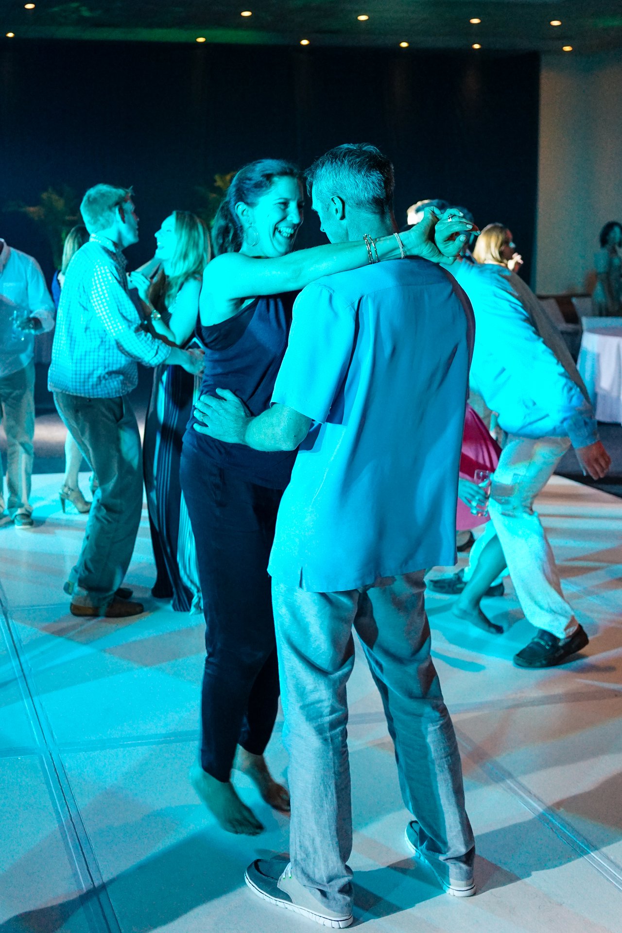 People dance and celebrate at an award party, with a smiling couple in the foreground enjoying the moment.