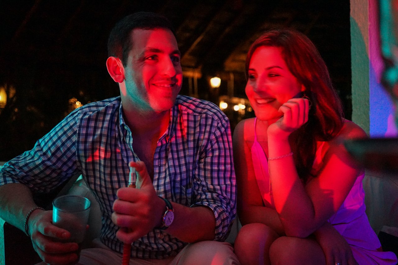 A man and woman sit together at a party, smiling and talking under colorful lighting.