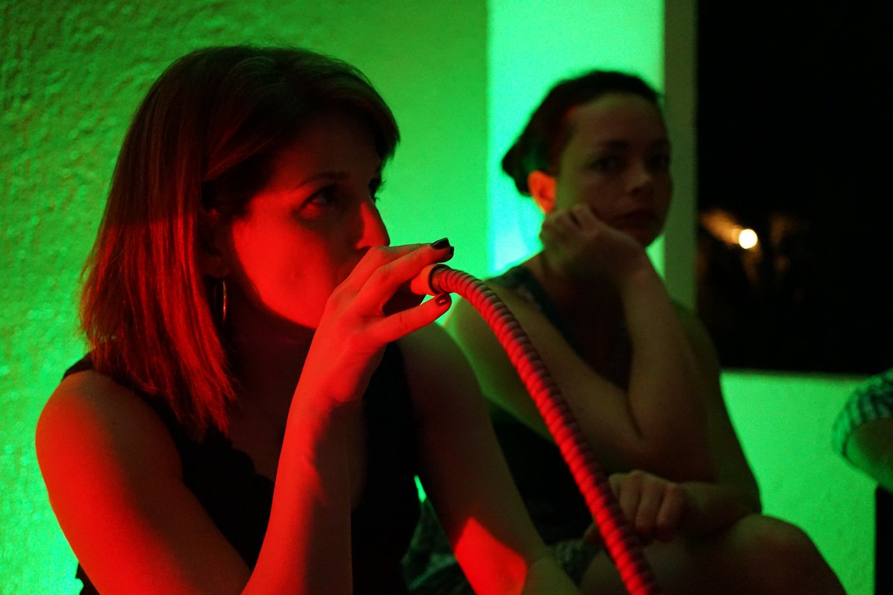 A woman smokes a hookah while another woman sits nearby, watching in a dimly lit party setting.