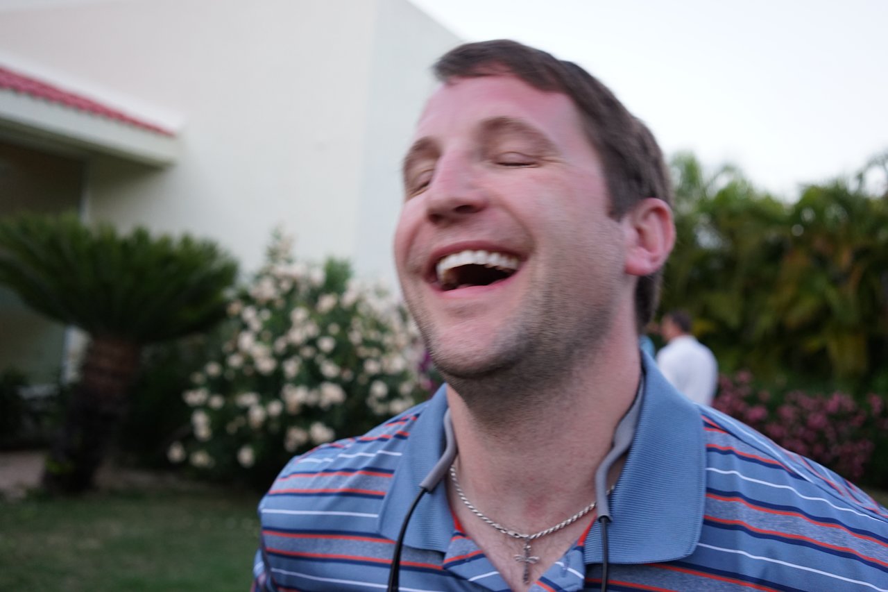 A man in a striped shirt laughs with his eyes closed at an outdoor award party.