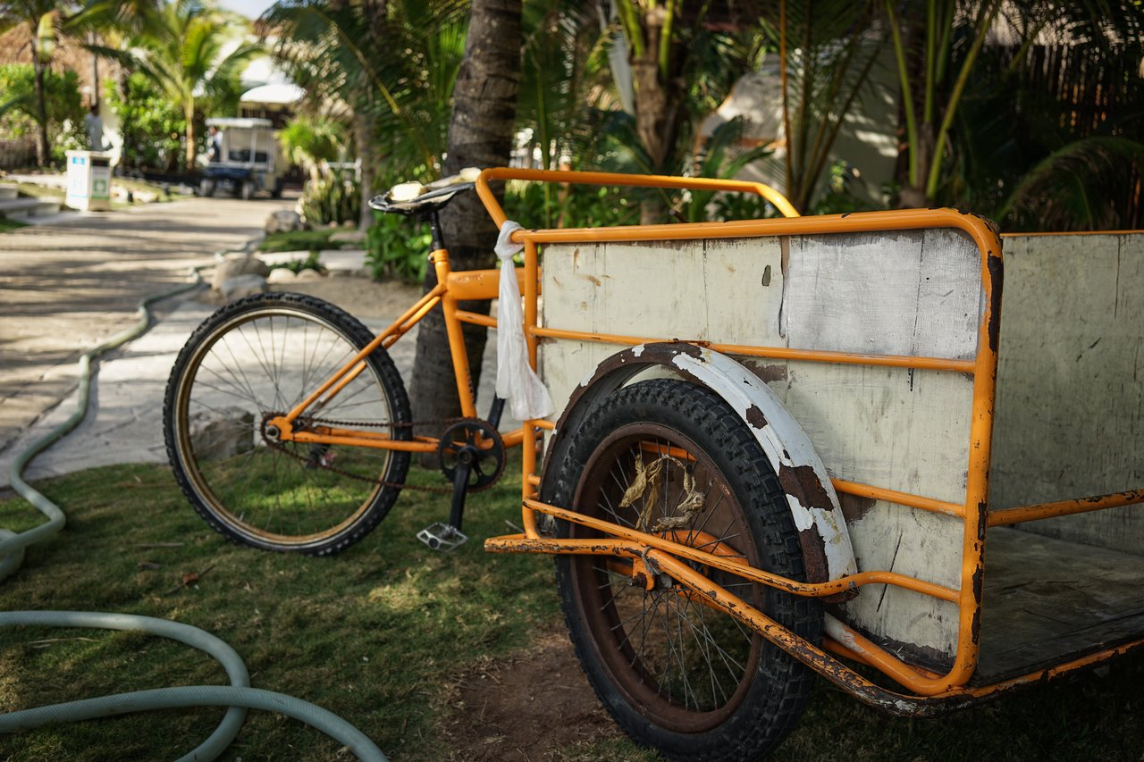 A yellow cargo tricycle with a large wooden cart is parked on grass near a tropical setting.