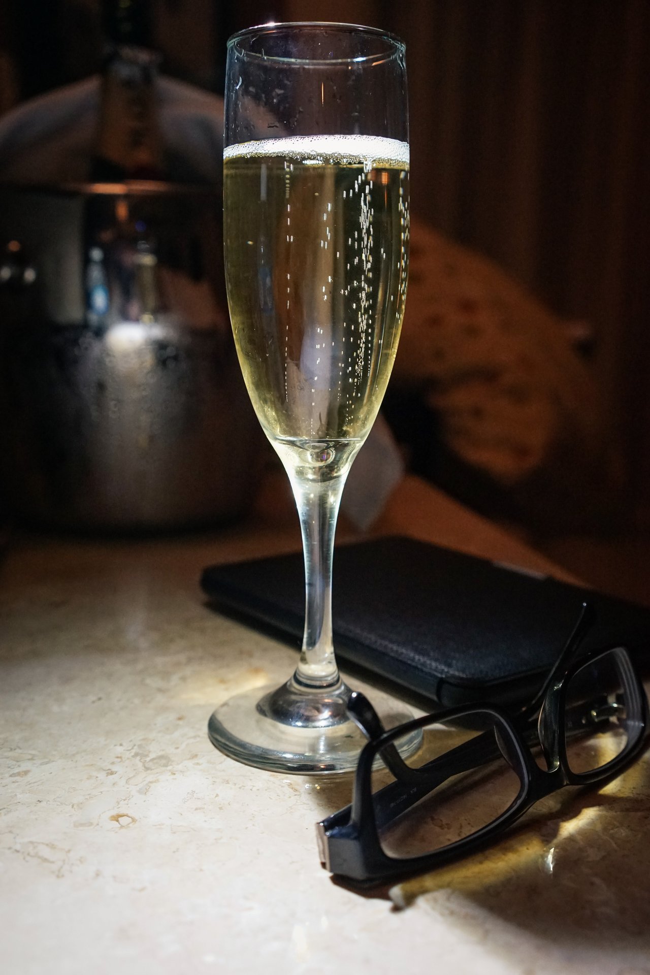 A glass of sparkling wine with bubbles rising, placed on a table next to glasses and a notebook.