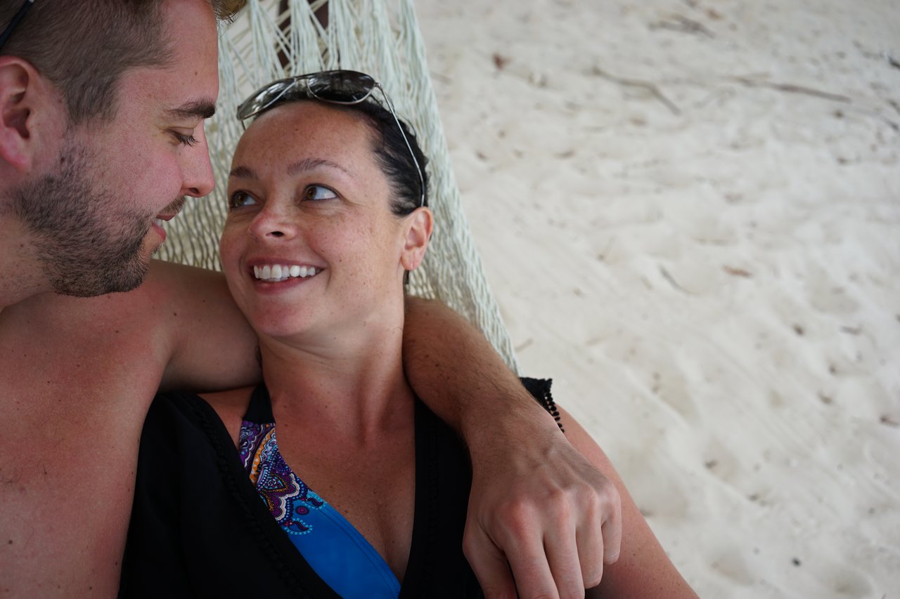 A couple relaxes in a hammock on a sandy beach, smiling and looking at each other.