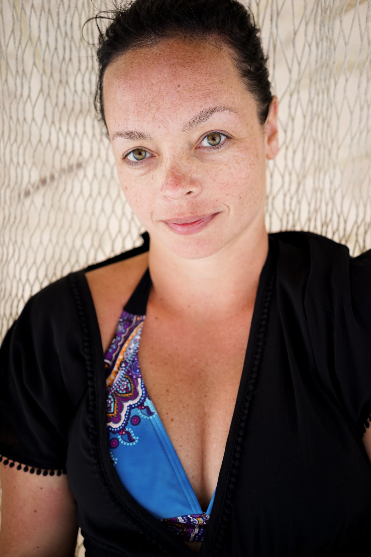 A woman in a black cover-up and blue swimsuit relaxes in a hammock, looking directly at the camera.