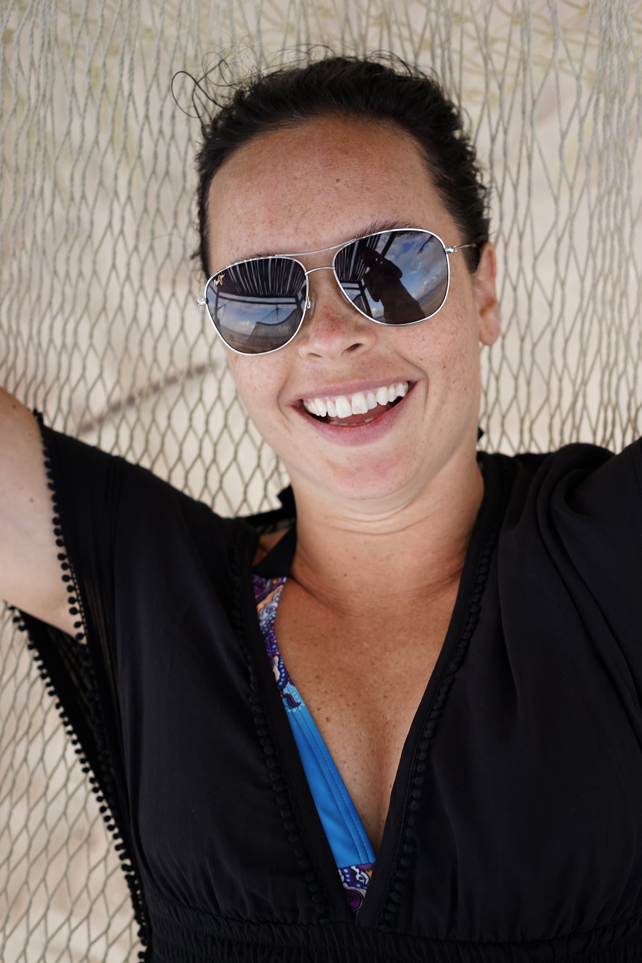 A smiling person wearing sunglasses and a black cover-up relaxes in a hammock, with a reflection in their lenses.