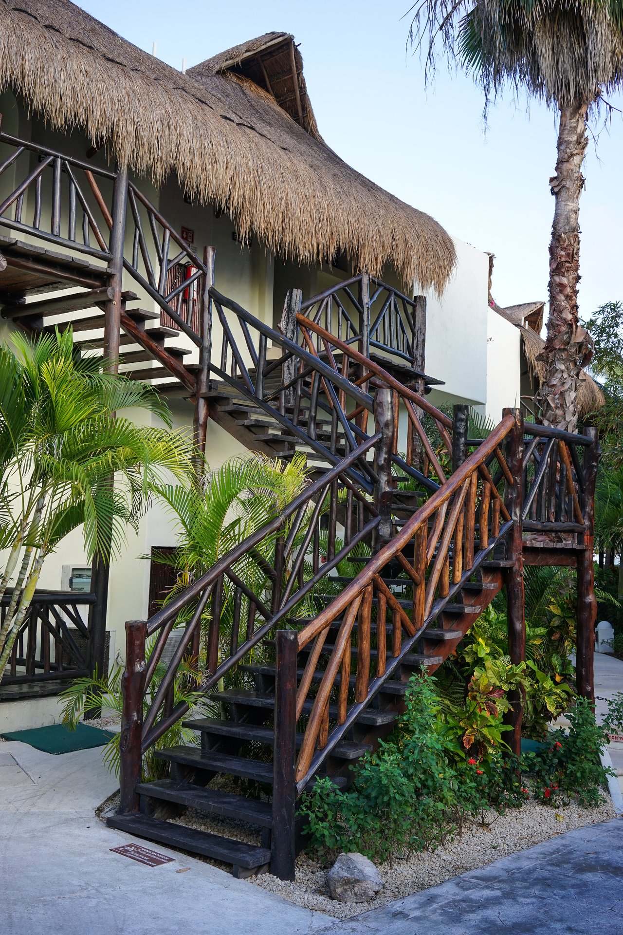 Thatched-roof hotel building with wooden stairs leading to upper rooms, surrounded by green plants and palm trees.