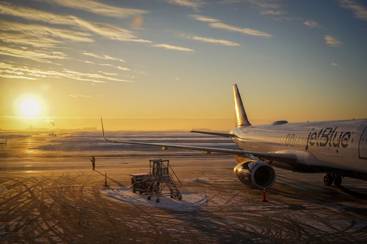 A JetBlue airplane is parked on a snowy tarmac at Logan Airport during sunrise.