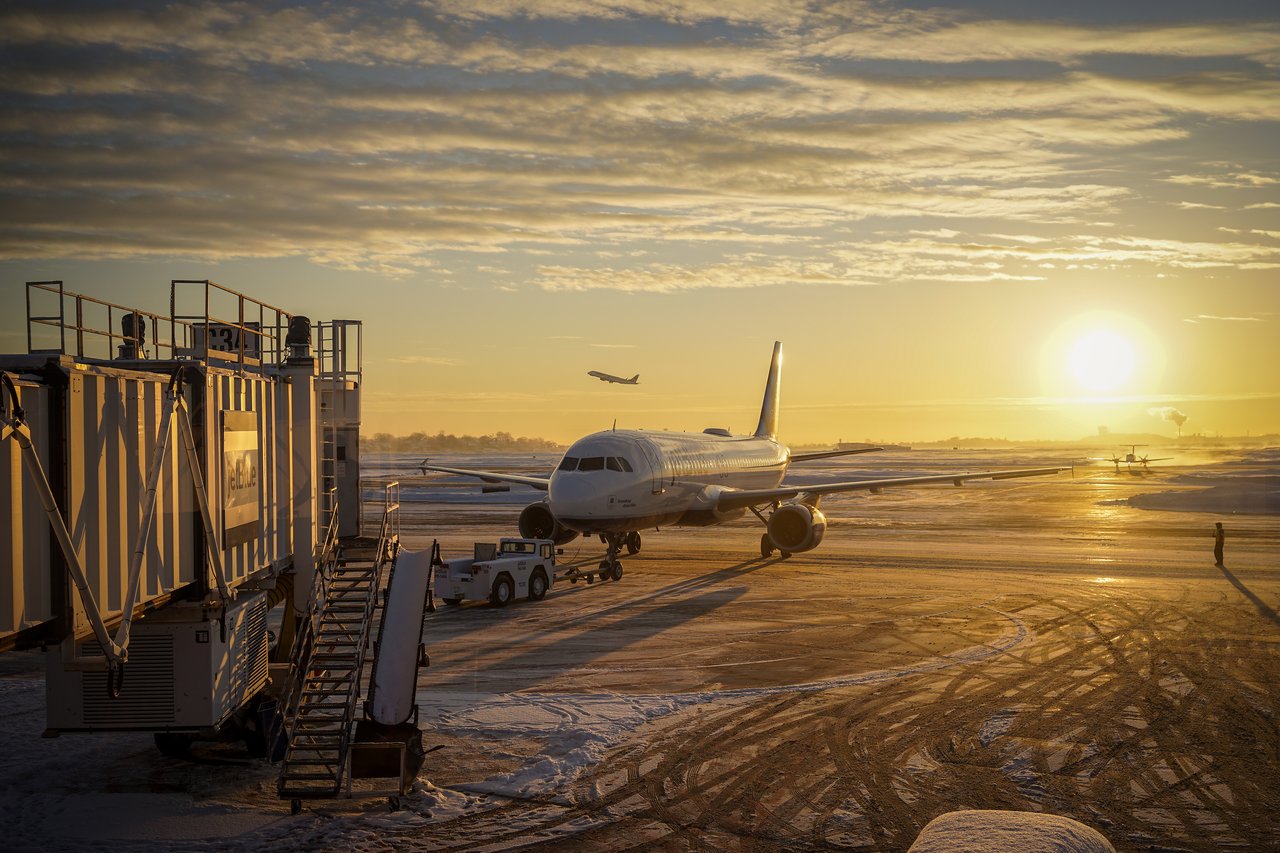 A JetBlue airplane is parked at Logan Airport near a jet bridge, with ground crew and equipment nearby.