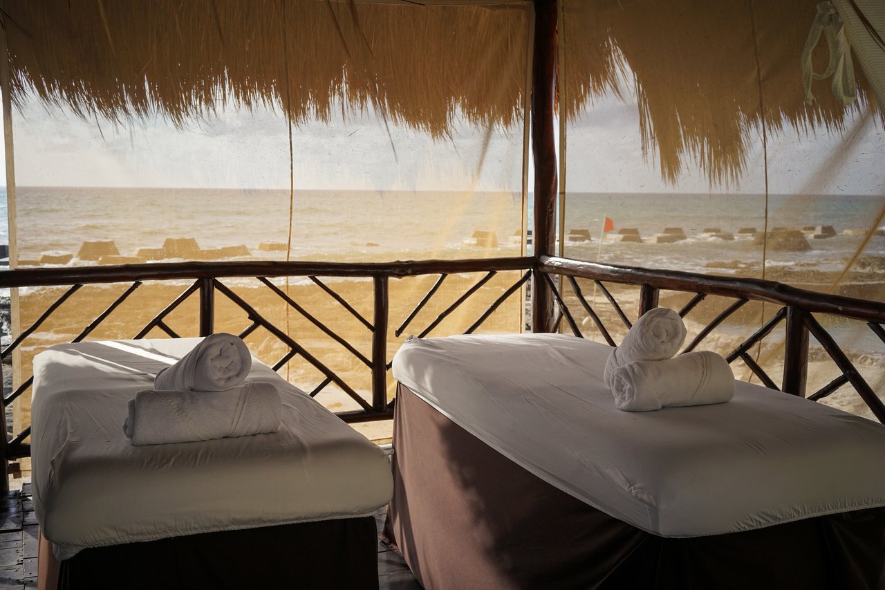 Two massage tables with rolled towels set up in a thatched-roof cabana overlooking the ocean.