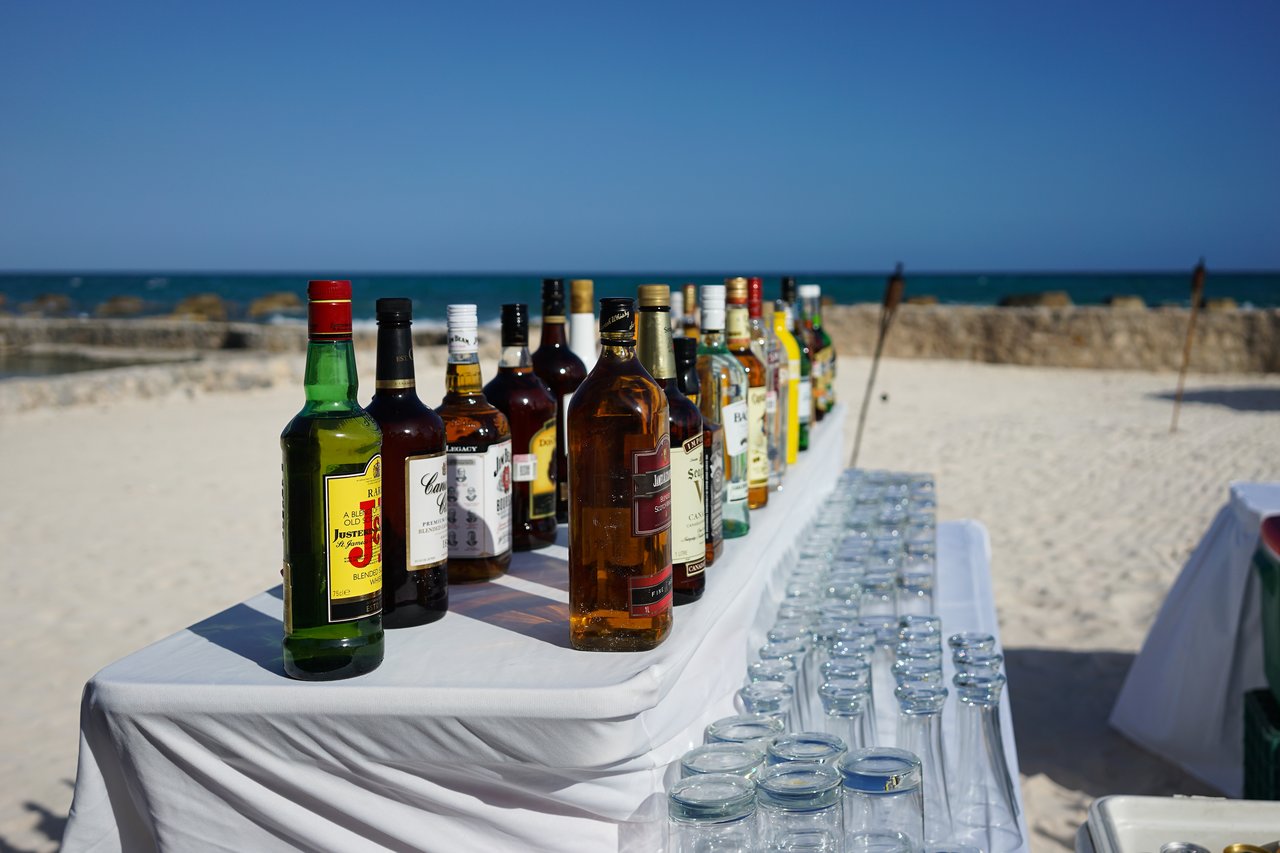 A beachside bar with liquor bottles and empty glasses arranged on a white table near the ocean.