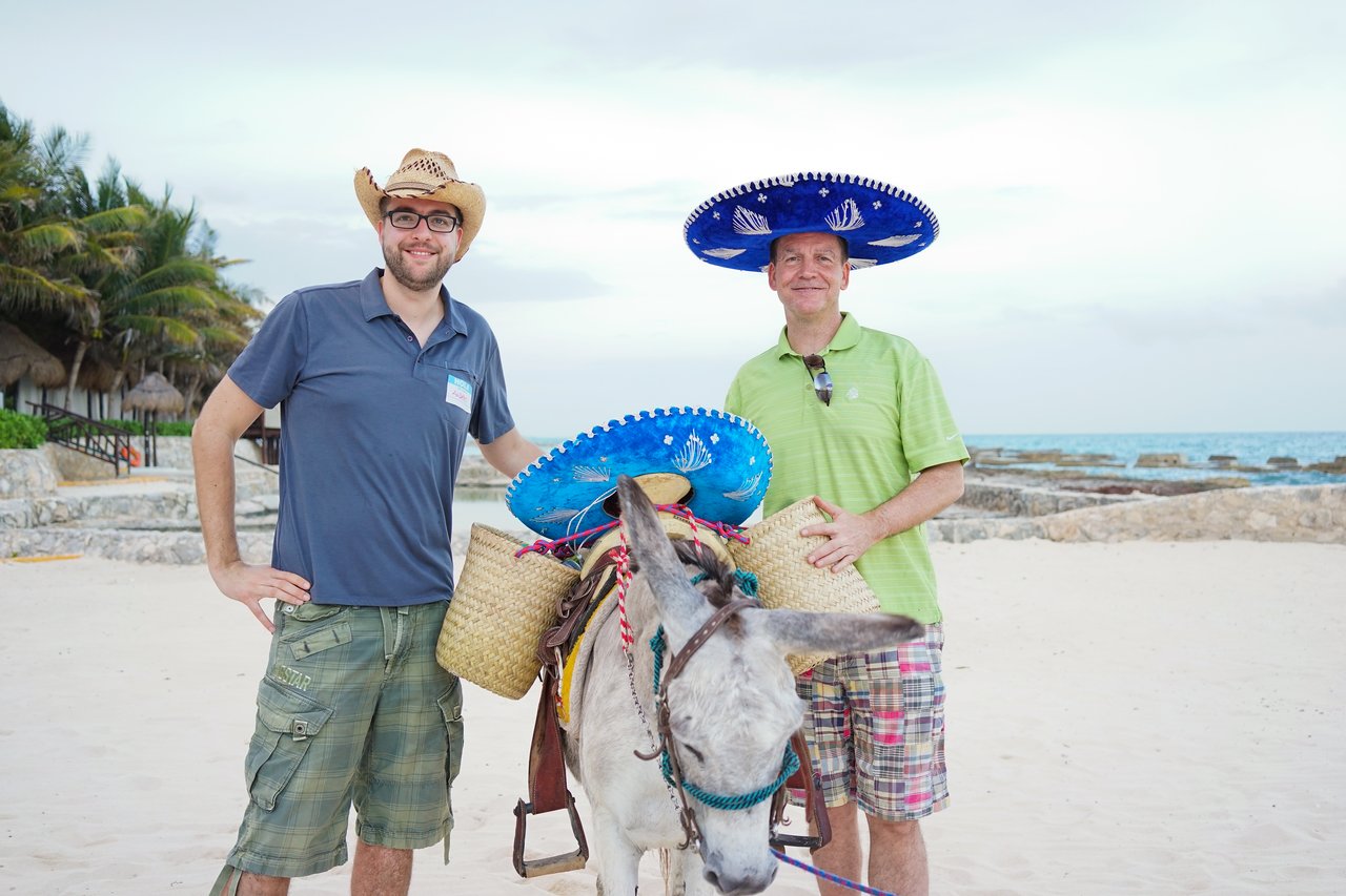 Two men wearing sombreros pose with a donkey carrying baskets on a sandy beach.
